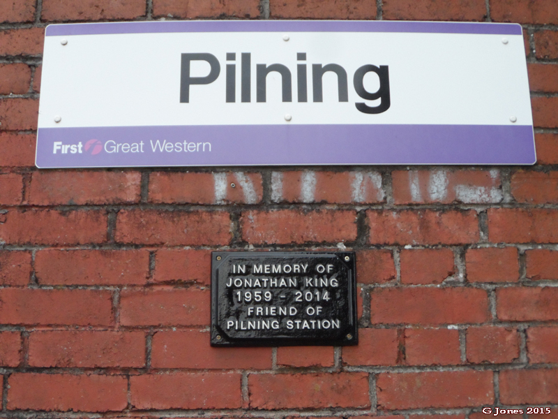 A picture, showing the Pilning running-in board branded First Great Western. Underneath is the memorial plaque commemorating Jonathan King. The plaque reads, "In memory of Jonathan King. 1959-2014. Friend of Pilning Station."